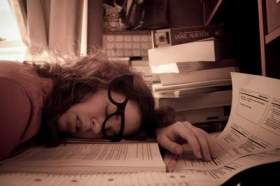 An exhausted woman falls asleep at her desk, amidst paper and notebooks.