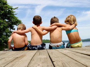 Four kids who are friends, sit on a dock with their arms around each others'' shoulders.